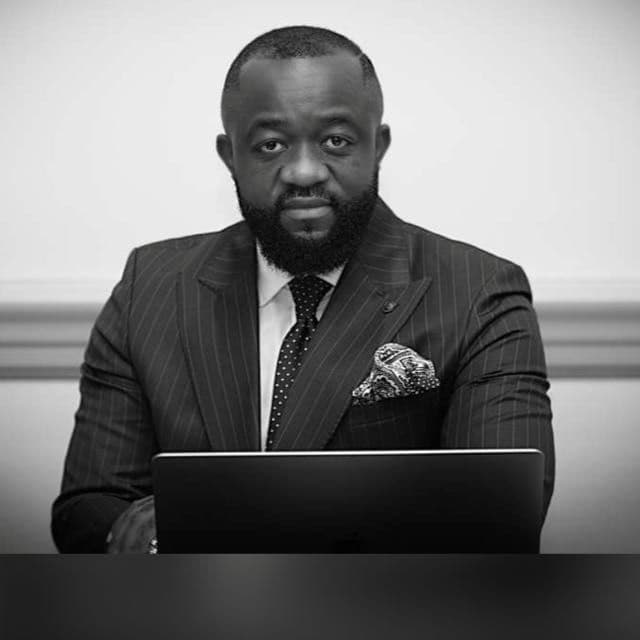 Bearded man in pinstripe suit and tie sitting behind a laptop in black and white.